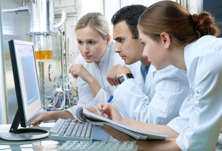 Three life sciences researchers in a lab facility looking at a report on a computer screen and taking notes