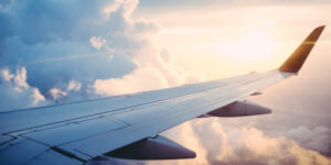 A view from an airplane window that shows its wing in a cloudy sky while the sun is setting in the background