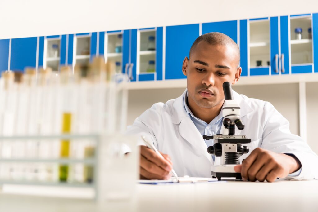 An individual sitting at a laboratory bench looking through a microscope and taking notes on a clipboard.